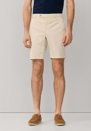 Homme portant un short beige au genou, une chemise bleu marine et des chaussures en daim marron sans lacets, debout devant un fond gris clair uni.