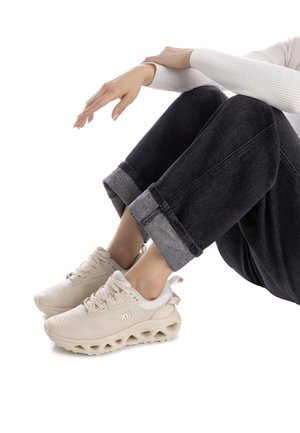 Zapatillas deportivas beige con parte superior de malla, suela gruesa y acolchada con detalles recortados, combinadas con pantalones de mezclilla negros enrollados.