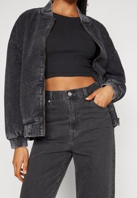 Veste bomber en denim noir à texture lisse, fermeture éclair et poignets côtelés, associée à un jean noir taille haute et un crop top ajusté.