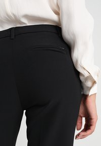 Mustat räätälöidyt housut, joissa on sileä pinta, ja joissa on pieni vetoketjullinen tasku takana sekä kapea malli.