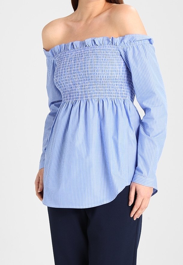 Femme portant un chemisier rayé bleu et blanc à épaules dénudées avec corsage smocké et manches longues, associé à un pantalon foncé.