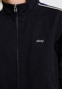 Melnā rāvējslēdzēja jaka ar augstu apkakli, ar smalku "obey" logotipu baltā krāsā. Materiāls šķiet tekstūrēts, ar sānu kabatām un kontrastējošām svītrām.