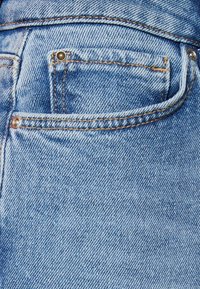 Jeans de mezclilla de color azul claro con un bolsillo trasero con un remache de metal, detalles de costura visibles y una textura suave.