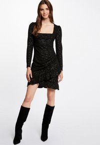 Robe noire à motifs avec un décolleté carré, des manches longues et un ourlet froncé. Associée à des bottes en suédine noires jusqu'aux genoux.