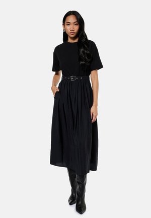 Vrouw met lang donker haar, gekleed in een zwarte midijurk met ceintuur en zwarte kniehoge laarzen, staand met één hand in haar zak.