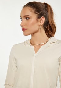 Felpa zip con cappuccio di un beige chiaro, caratterizzata da una texture liscia, abbinata a collane di metallo dorato a strati e orecchini a cerchio.