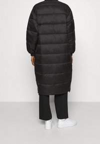 Sort polstret lang frakke med quiltet design, elastiske manchetter og sidelommer med lynlås. Sammen med sorte bukser og hvide sneakers.