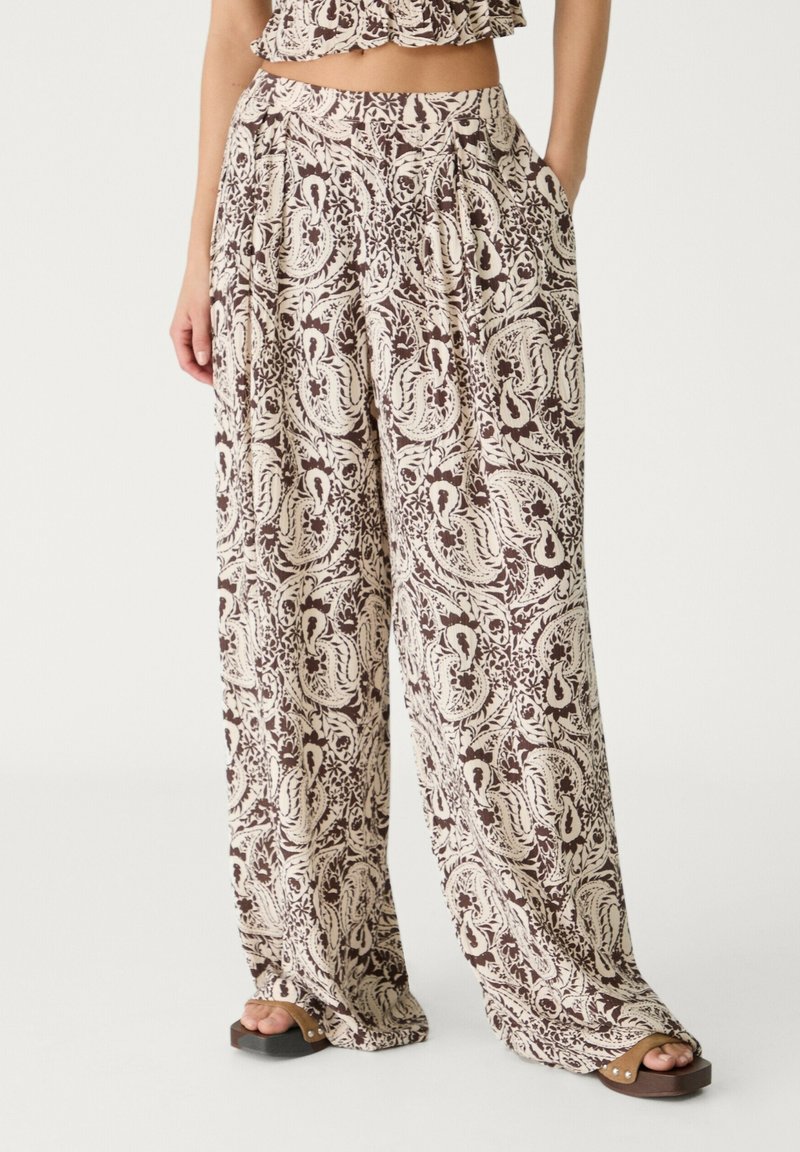 Vrouw in wijde broek met bruin en crème paisleypatroon, gecombineerd met bruine plateau-sandalen op een witte achtergrond.