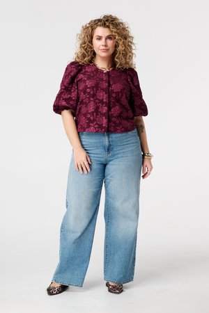 Krullige vrouw met een paarse blouse met bloemenprint en pofmouwen, lichtblauwe wijde jeans, luipaardprint schoenen, staand tegen een witte achtergrond.