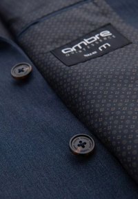 Granatowa koszula z tkaniny w wzór w jodełkę, z kontrastową kieszenią w geometryczny wzór i ciemnobrązowymi guzikami. Wyeksponowana metka marki.