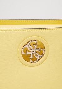 Żółta skórzana torebka z okrągłym złotym logo na środku. Charakteryzuje się kontrastującymi przeszyciami i gładką fakturą.