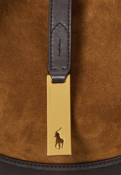 Geantă din suede maro cu o curea din piele închisă la culoare, având un tag rectangular auriu embossat cu un logo de cal. Textură netedă și design modern.