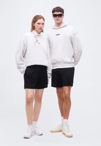 Δυο μοντέλα στέκονται δίπλα-δίπλα φορώντας ανοιχτό γκρι υπερμεγέθη φούτερ, μαύρες βερμούδες, λευκές κάλτσες και αθλητικά παπούτσια, το ένα με γυαλιά ηλίου και φουλάρι.