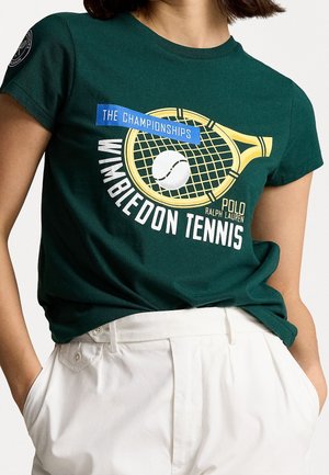Ciemnozielona koszulka tenisowa Wimbledon z żółtą grafiką rakiety i piłki tenisowej, zestawiona z białymi spodniami z wysokim stanem, ręce w kieszeniach.