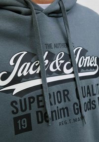Grå hættejakke lavet af blødt materiale, med et stort sort-hvidt grafisk logo med tekst, snorehætte og ribbede detaljer.