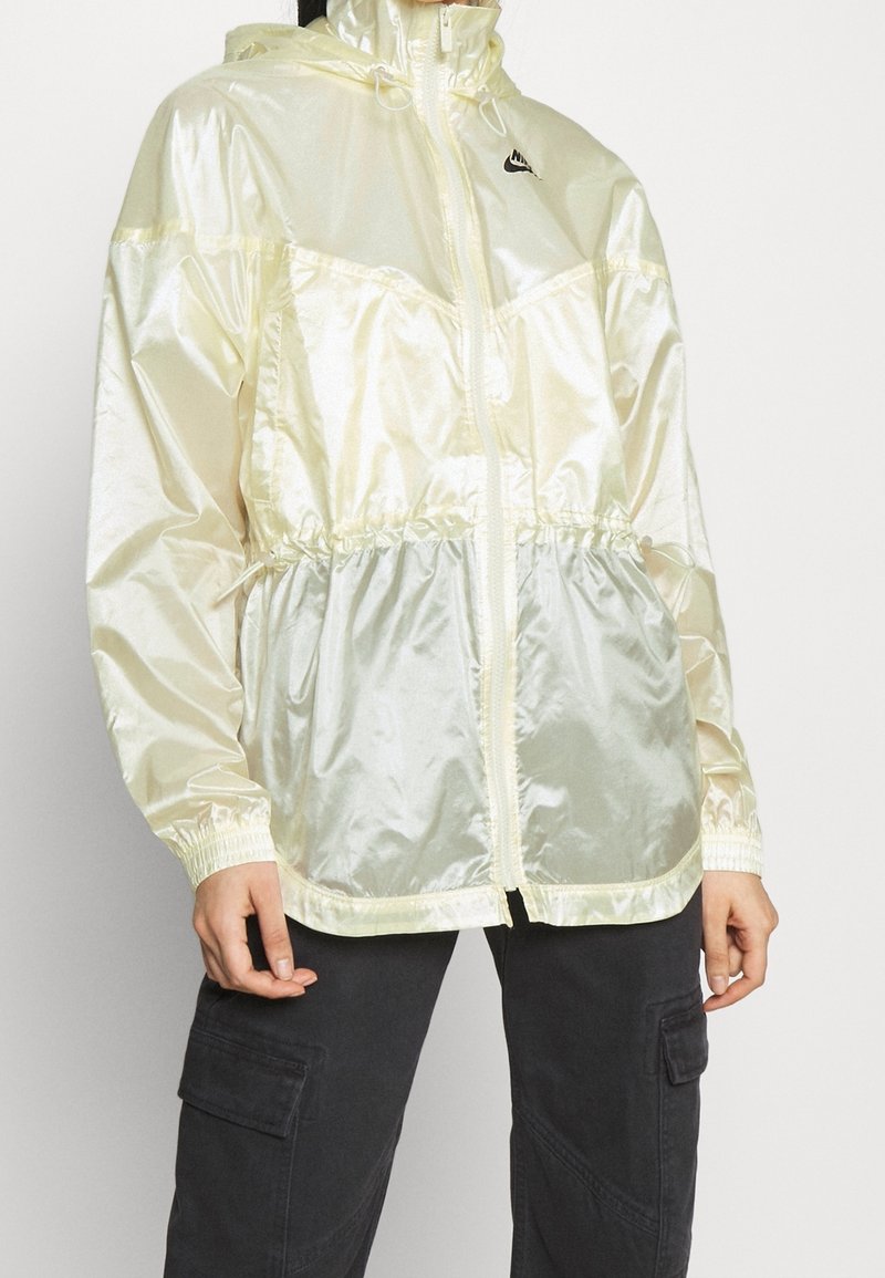 Ljust gul, glänsande nylonjacka med huva, elastisk midja och ärmslut. Har en framsida med dragkedja och svart logotyp på bröstet.