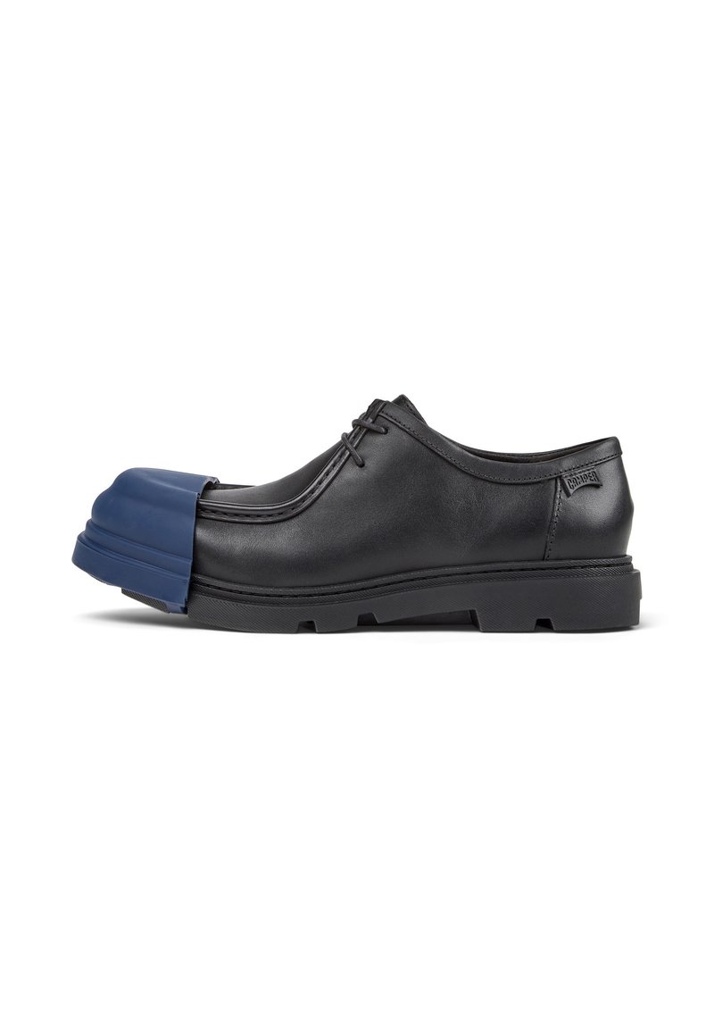 Zapato negro de cordones con una exclusiva puntera de goma azul, que presenta una suela texturizada y un diseño minimalista, exhibido sobre un fondo blanco.
