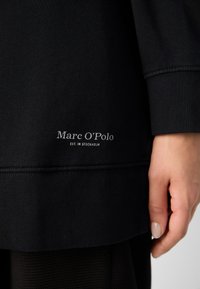 Gros plan d'un vêtement noir avec le texte "Marc O'Polo EST. IN STOCKHOLM" brodé près de l'ourlet et la main d'une personne reposant à côté.