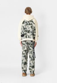 Sudadera con capucha en color crema con un patrón de mármol en verde, negro y blanco; combinada con pantalones a juego que presentan una marca y un bolsillo lateral.