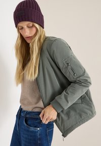 Olivenfarvet bomberjakke med lynlås foran og ribbede manchetter, båret over en beige sweater, parret med blå denimjeans og en lilla beanie.