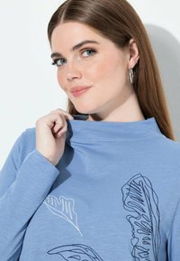 Ljusblå långärmad topp med en mock neck, med svart och vit bladbroderi. Slen yta och figurnära silhuett.