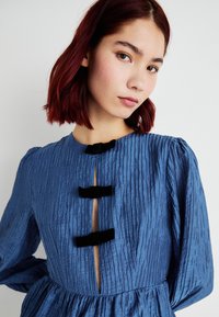 Vestido azul plissado com decote redondo, três detalhes em laço de veludo e um recorte no peito. Mangas longas conferem volume.