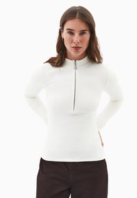 Mujer con cabello castaño, vistiendo pendientes de aro dorados, una camiseta de manga larga de canalé blanca con cremallera frontal y pantalones oscuros, de frente.