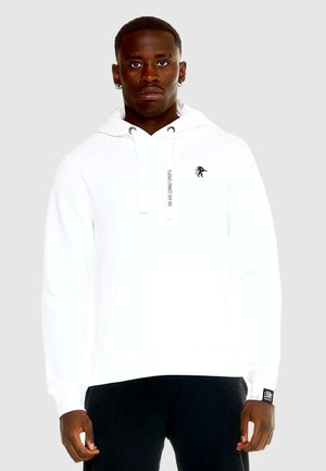 Homme portant un sweat à capuche blanc avec poche avant et logo lion noir, associé à un pantalon noir sur un fond uni.