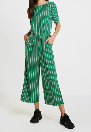 Grün gestreifter Overall mit kurzen Ärmeln und elastischem Bund, ausgestattet mit Seitentaschen und einem weiten Bein. Kombiniert mit schwarzen Sneakers.