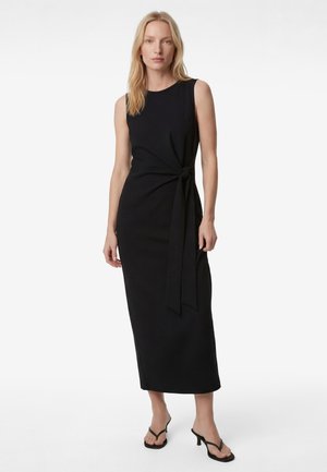 Femme portant une robe midi noire sans manches avec un nœud sur le côté et des sandales à talons noires, posant devant un fond blanc uni.