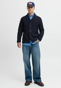 Marine gebreide cardigan met kabeldetails, vijf knopen, gelaagd over een denim shirt en gecombineerd met een losse blauwe jeans en bruine laarzen.