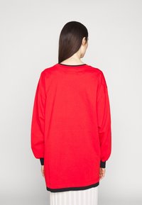 Kvinna med långt mörkt hår, iklädd en lös röd tröja med svarta muddar och fåll, som står mot en enkel ljus bakgrund och vänder ryggen mot kameran.