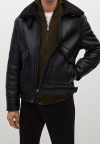 Giacca di pelle nera con colletto in shearling, chiusura con zip, due tasche laterali e interno di un verde oliva testurizzato, indossata sopra pantaloni scuri.