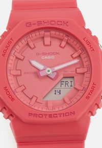 Roza G-SHOCK ura z okroglo digitalno zasnovo, plastično ohišje in teksturiranim trakom. Vključuje večnamenske gumbe in prikaz datuma.