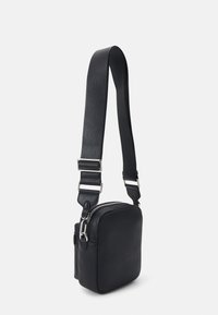 Crna kožna torba preko tijela kvadratnog oblika, s podesivim remenom, srebrnim detaljima i zatvaranjem na patentni zatvarač. Glatka tekstura i minimalistički dizajn.