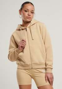 Felpa con zip beige e cappuccio, con coulisse, tasca frontale e logo bianco ricamato. Indossata con pantaloni corti beige coordinati. Texture liscia.