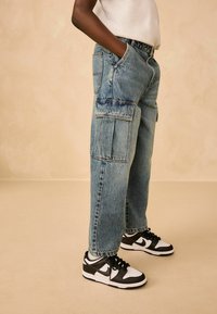 Jeans cargo lejere, de culoare albastru deschis, cu două buzunare laterale mari, margini destrămate și cusături în contrast, asortați cu adidași negri și albi.