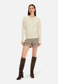 Cremfarbener Strickpullover mit geripptem Saum, kombiniert mit gemusterten braunen Shorts und kniehohen braunen Wildlederstiefeln. Stehende Pose, neutraler Hintergrund.