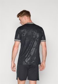 Sort t-shirt med korte ærmer og blomstret mønster, med en stribet krave. Bæret med mørke shorts, hvilket viser en afslappet atletisk stil.