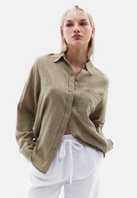 Olivově zelená košile na knoflíky s texturovanou látkou, mírně oversize střih, a náprsní kapsou, doplněná bílými kalhotami s tónovaným stahováním.