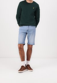 Grön sweatshirt med rund hals och en liten gul logga, matchad med ljusblå denimshorts. Bruna sneakers och vita strumpor fulländar outfiten.