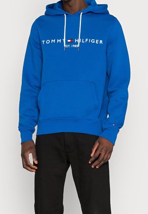 Osoba nosząca niebieską bluzę z kapturem Tommy Hilfiger z białymi sznurkami i czarne spodnie na jednolitym jasnym tle.