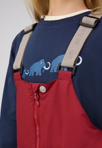 Rode overalls met een ritssluiting en knopen, voorzien van beige bandjes. Het blauwe sweatshirt eronder heeft een afbeelding van olifanten.