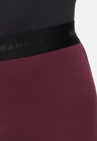 Nahaufnahme eines schwarzen elastischen Taillenbunds mit dem Text "MAMMUT" auf weinrotem Stoff, getragen über dunklem geripptem Material.