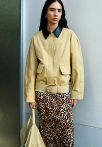 Beige Jacke mit schwarzem Kragen und Fronttaschen, kombiniert mit einem Rock im Leopardenmuster. Hält eine beigefarbene Tasche. Glatte Textur und strukturiertes Design.