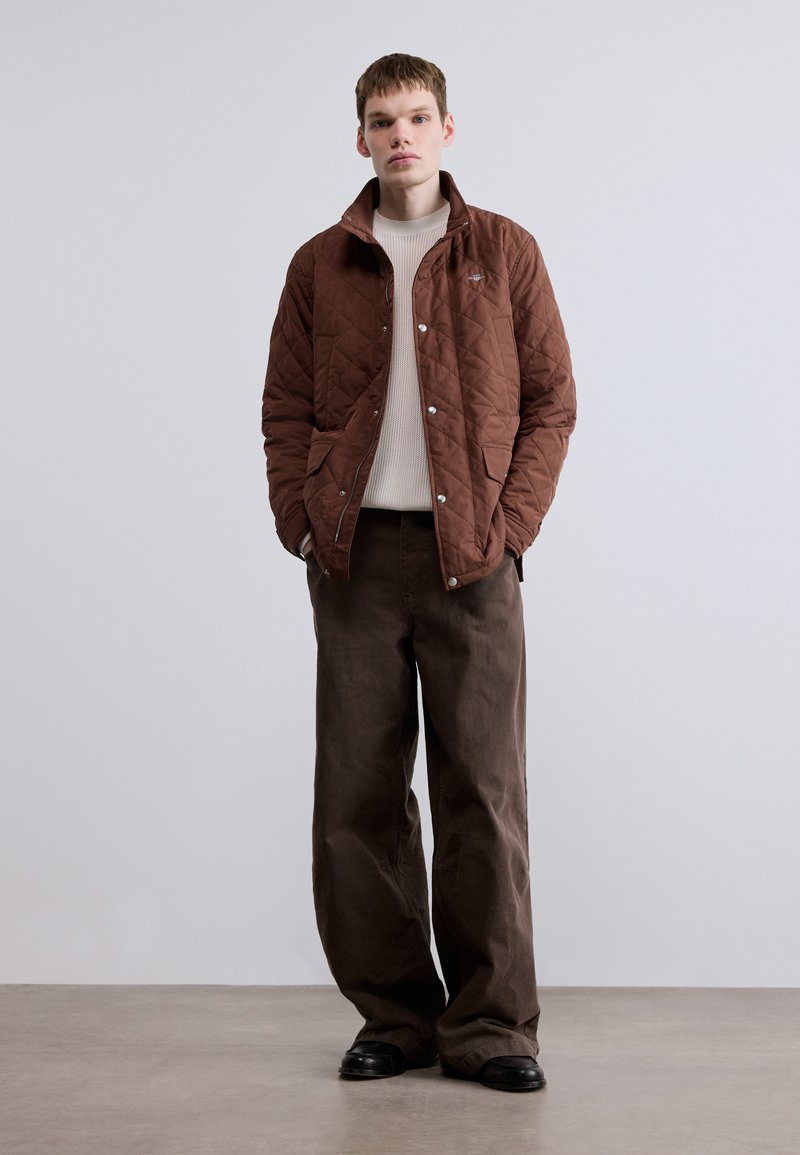 Veste matelassée marron avec un col montant et des boutons-pression. Portée sur un pull en maille crème, associée à un pantalon large marron et des chaussures noires.