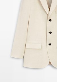 Beżowa marynarka z czarnymi guzikami i przednią kieszenią z klapką, pokazana od talii w dół na białym tle.