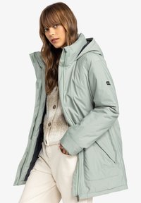 Jachetă puffer de culoare verde deschis, cu o glugă mare, fermoar frontal și buzunare laterale. Prezintă un căptușeală neagră și elemente integrate ajustabile.