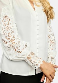 Biela blúzka s čipkovanými rukávmi a gombíkmi potiahnutými látkou, zobrazujúca zložené ruky s prsteňmi a umením na nechtoch na svetlej pokožke.