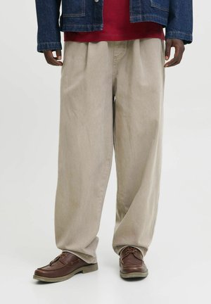 Mand iført løstsiddende beige bukser, brune lædersko, rød skjorte og en blå denimjakke, stående foran en ensfarvet baggrund.